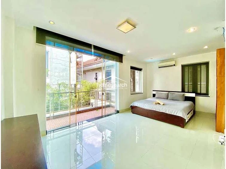 This minimalist bedroom is finished with light-colored tile flooring and white walls, emphasizing the sense of space and natural light provided by the large sliding glass doors. The room is furnished with a simple bed and a dark wood headboard, offering a clean, uncluttered aesthetic that connects seamlessly to the outdoor balcony.