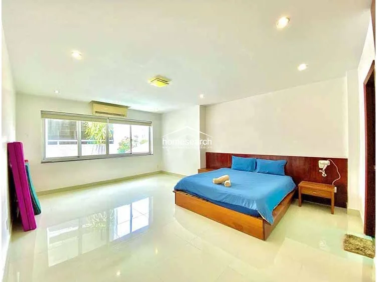 This bright bedroom maintains a consistent design theme with neutral walls, polished tile floors, and a wall-mounted AC unit for climate control. A large window provides an expansive view, while the room is furnished with a comfortable bed, a wooden headboard, and a matching bedside table, presenting a well-maintained and orderly living space.