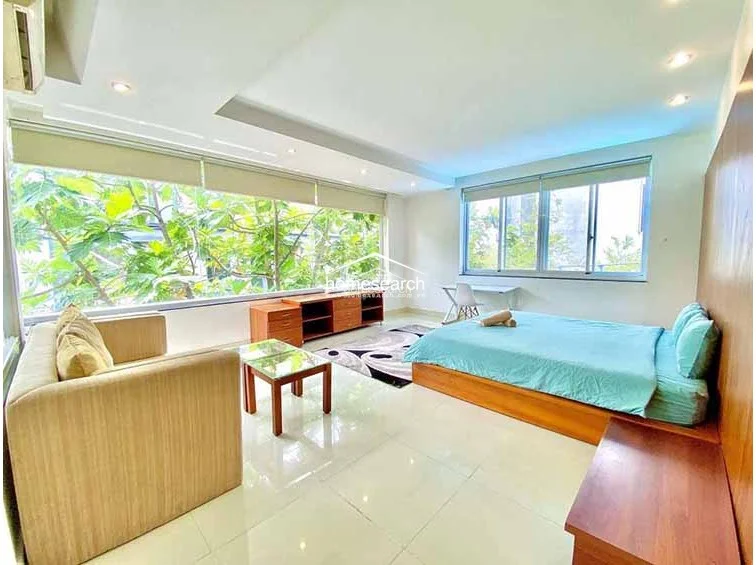 This spacious bedroom features an open-concept layout with a dedicated lounge area, highlighted by expansive floor-to-ceiling windows that offer lush, green views and abundant natural light. The room is finished with light-colored tile flooring, recessed ceiling lighting, and a neutral white color scheme, complemented by a wooden bed frame and matching media console.