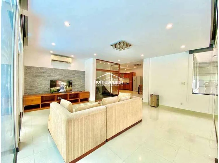 This spacious open-plan living area features glossy tile flooring and a neutral color palette, creating a bright and airy atmosphere. The room is anchored by a large beige sectional sofa facing a wall-mounted TV on a textured stone feature wall, with a clear flow leading directly into the dining and kitchen areas.