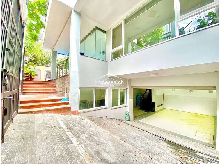 This exterior view shows a multi-level property with a paved driveway leading to a spacious, open-air garage area on the lower level. The structure features modern architectural lines, large glass windows for natural light, and a set of tiled stairs leading up to an elevated terrace or entrance, indicating a well-maintained, contemporary design.