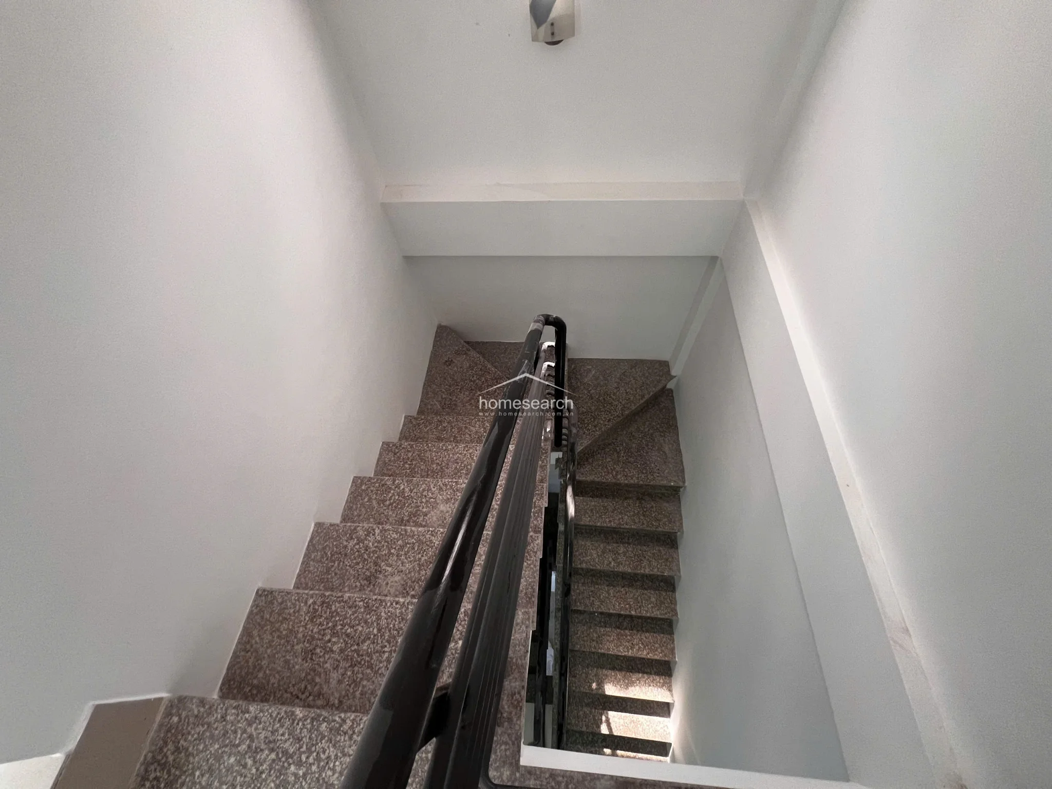 This view looking down the staircase highlights the durable, speckled stone-tiled steps and the sturdy black metal handrail. The stairwell is minimalist and clean, featuring stark white walls and a simple ceiling light fixture that maintains a bright, airy atmosphere throughout the transition between floors.