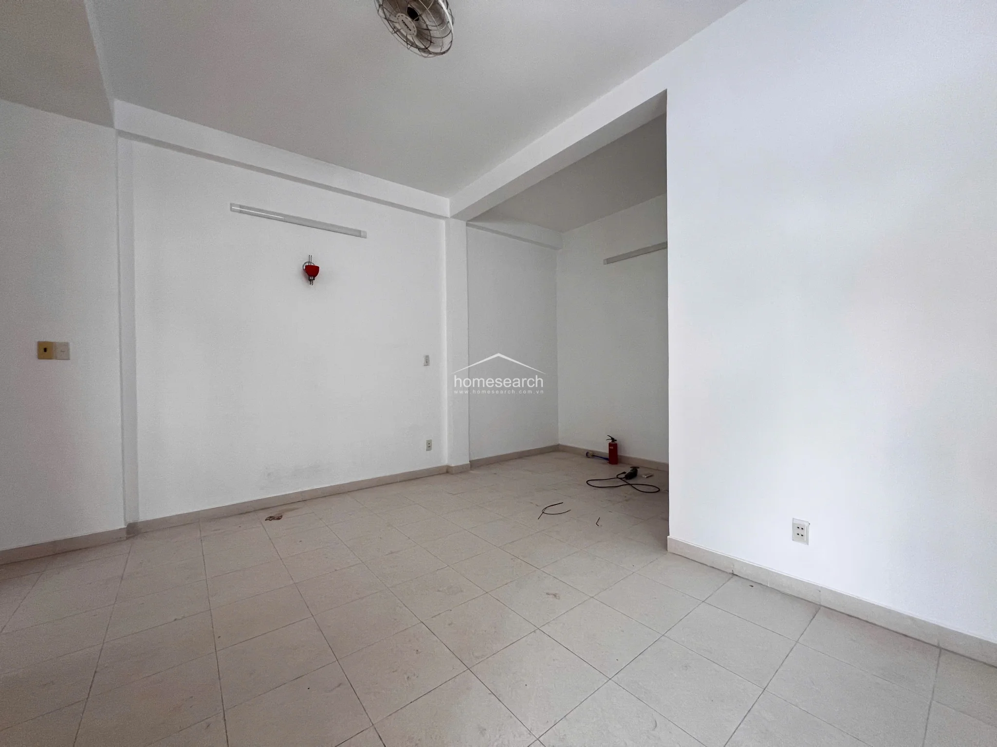 This versatile living space features a clean, minimalist aesthetic with white walls and light tile flooring. It includes a ceiling fan for climate control and a small red decorative accent, with an open layout that flows into a secondary alcove area.