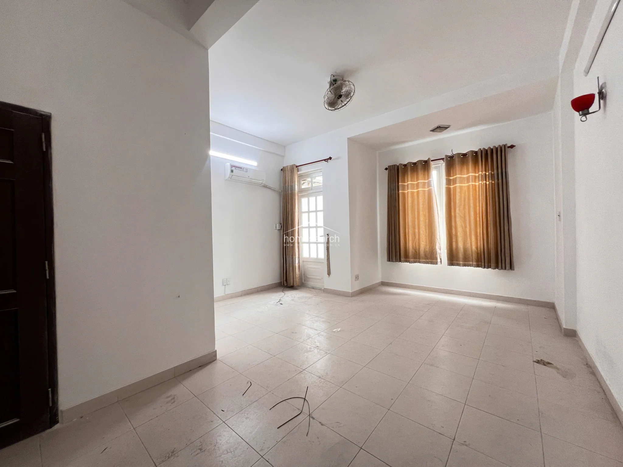 This bright, open-plan room features neutral white walls and light-colored floor tiles, creating a clean and minimalist aesthetic. It is equipped with a wall-mounted AC unit, a ceiling-mounted fan, and large windows dressed with warm-toned curtains that allow for ample natural light.