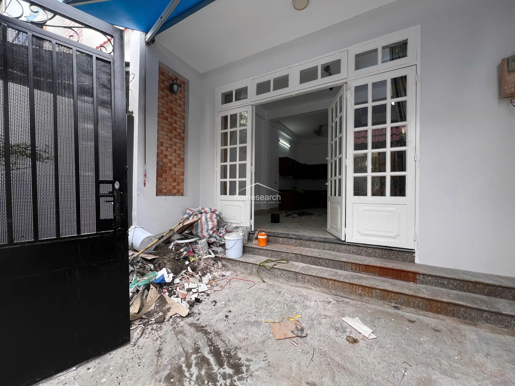 The exterior threshold displays a transition from the gated courtyard to the main interior, currently undergoing renovation as evidenced by construction debris. The entrance features wide, multi-paned white French doors and stone steps, with a decorative brick accent wall adding texture to the otherwise minimalist white facade.