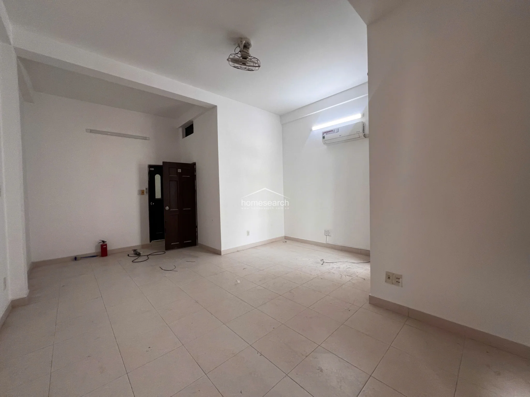 This open-concept living area boasts high ceilings and a neutral color palette, creating a bright and airy atmosphere. The space includes a wall-mounted air conditioning unit, a ceiling fan, and a dark wooden door, with tile flooring throughout for easy maintenance.