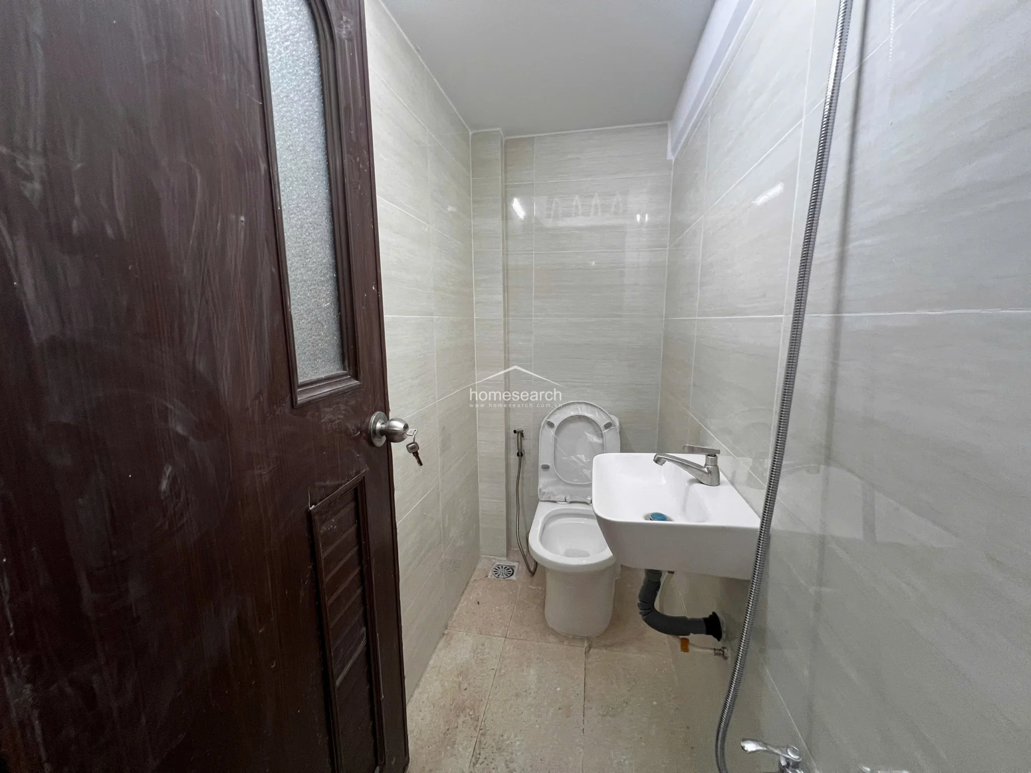 This compact bathroom is finished with light-colored wall and floor tiles, providing a neutral and easy-to-clean environment. It includes a standard white pedestal sink, a toilet, and a handheld bidet sprayer, though the exposed plumbing under the sink suggests the space is in basic, functional condition.