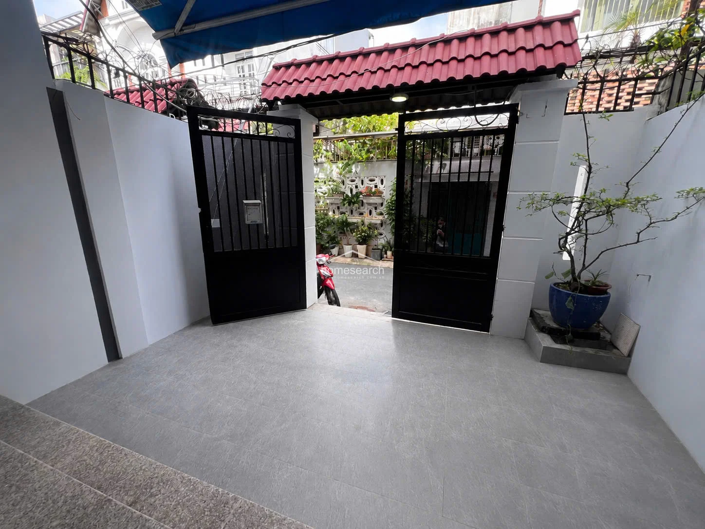This image shows a secure, gated entrance area featuring a neutral grey tiled floor and clean white walls. The space is framed by a traditional red-tiled roof structure over the black metal gates, with a potted plant adding a touch of greenery to the corner.