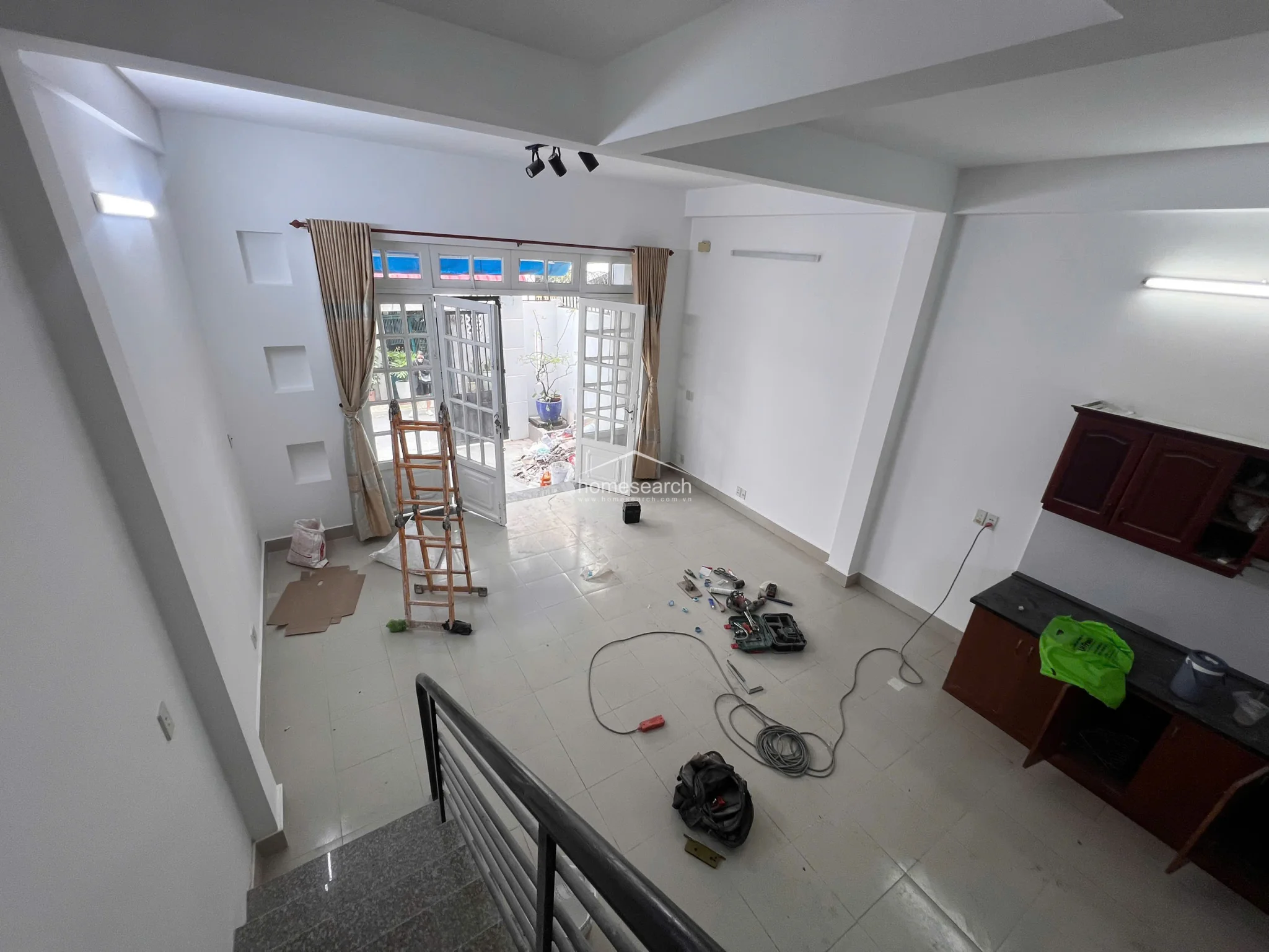 This wide-angle shot captures the open-concept ground floor, which integrates a living area and kitchen space under high ceilings. The room is currently in a state of active maintenance, featuring light-colored tile flooring, white walls, and a set of wooden kitchen cabinets, all illuminated by a combination of natural light from the entrance and overhead fluorescent lighting.
