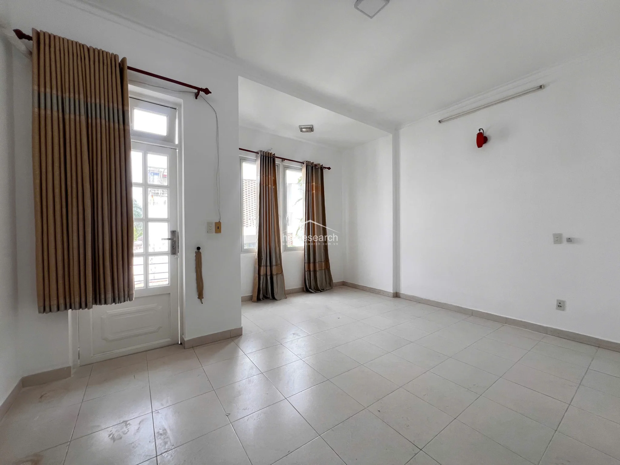 This bright, airy room is characterized by large windows and a glass-paned door draped with elegant brown curtains, allowing for ample natural light and a view of the outdoors. The space is finished with neutral tile flooring and white walls, featuring a minimalist design that includes a wall-mounted fire extinguisher and basic electrical fixtures.