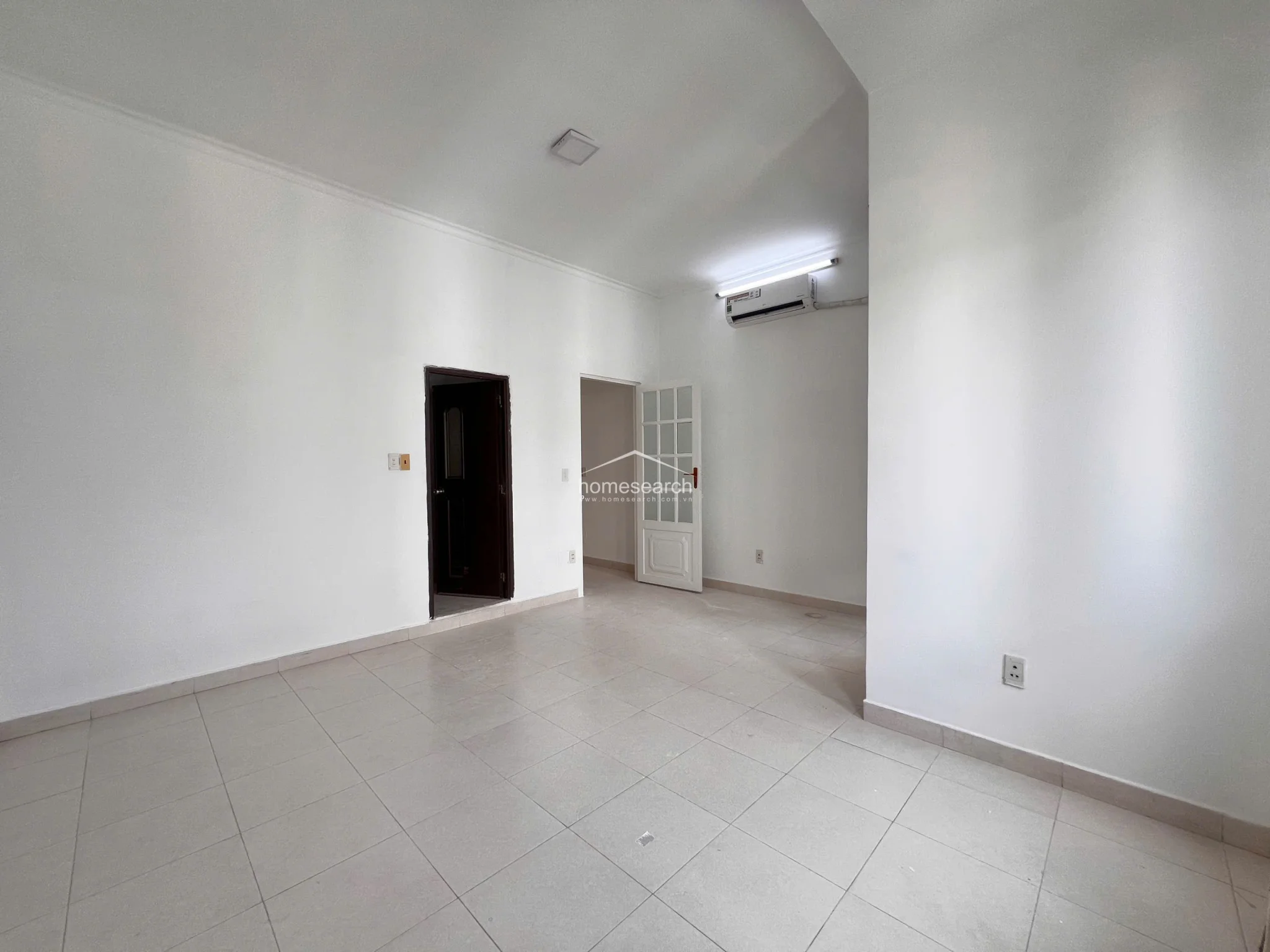 This spacious, unfurnished room features clean white walls and light tile flooring, offering a versatile blank canvas for any interior design style. The room is equipped with a wall-mounted air conditioning unit and features a white paneled door, providing a bright and well-maintained environment ready for immediate occupancy.