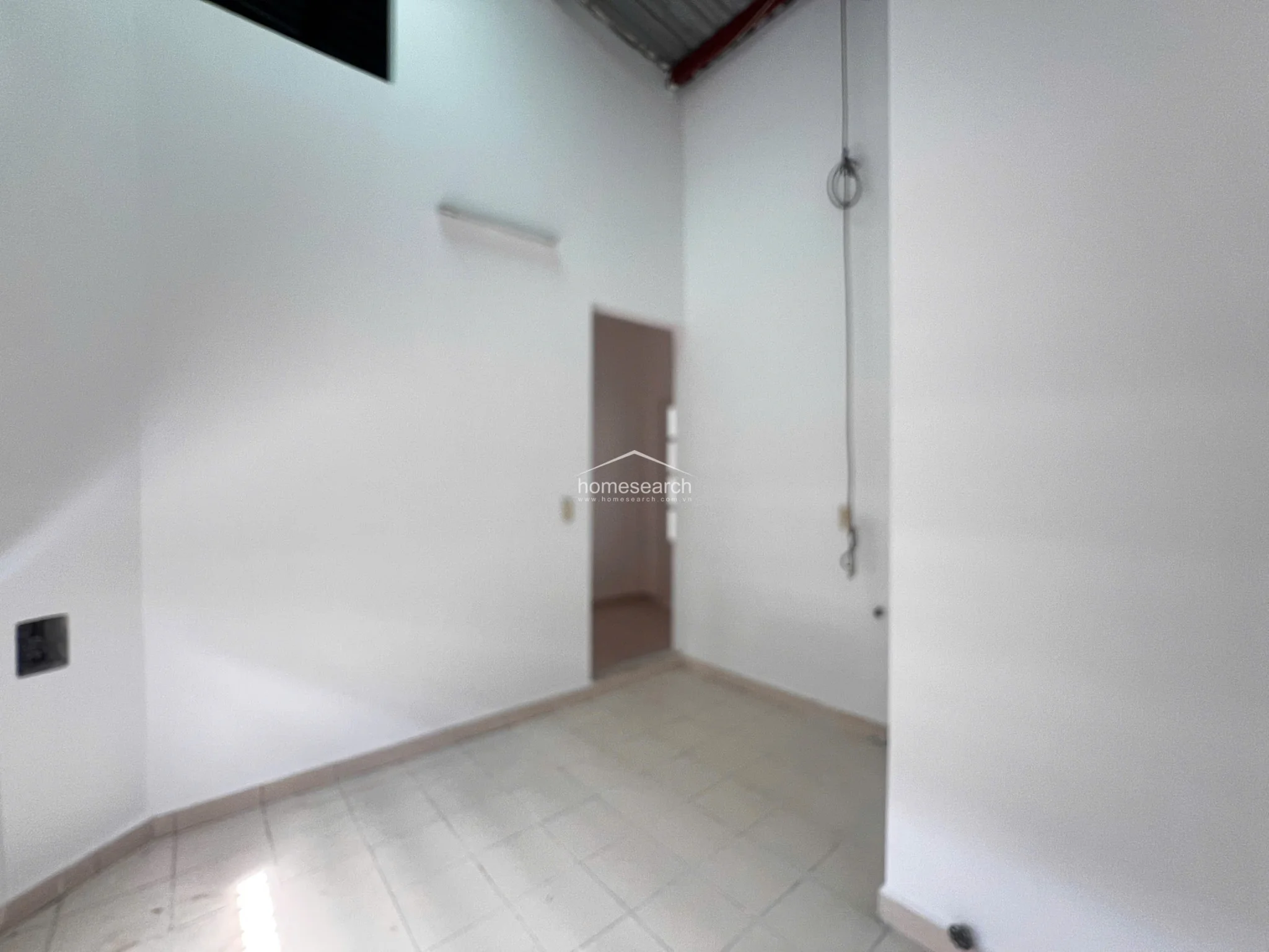 This image shows a minimalist, high-ceilinged room with neutral white walls and light-colored floor tiles. The space features a doorway leading to an adjacent area and includes basic electrical wiring, suggesting a functional, utilitarian layout.