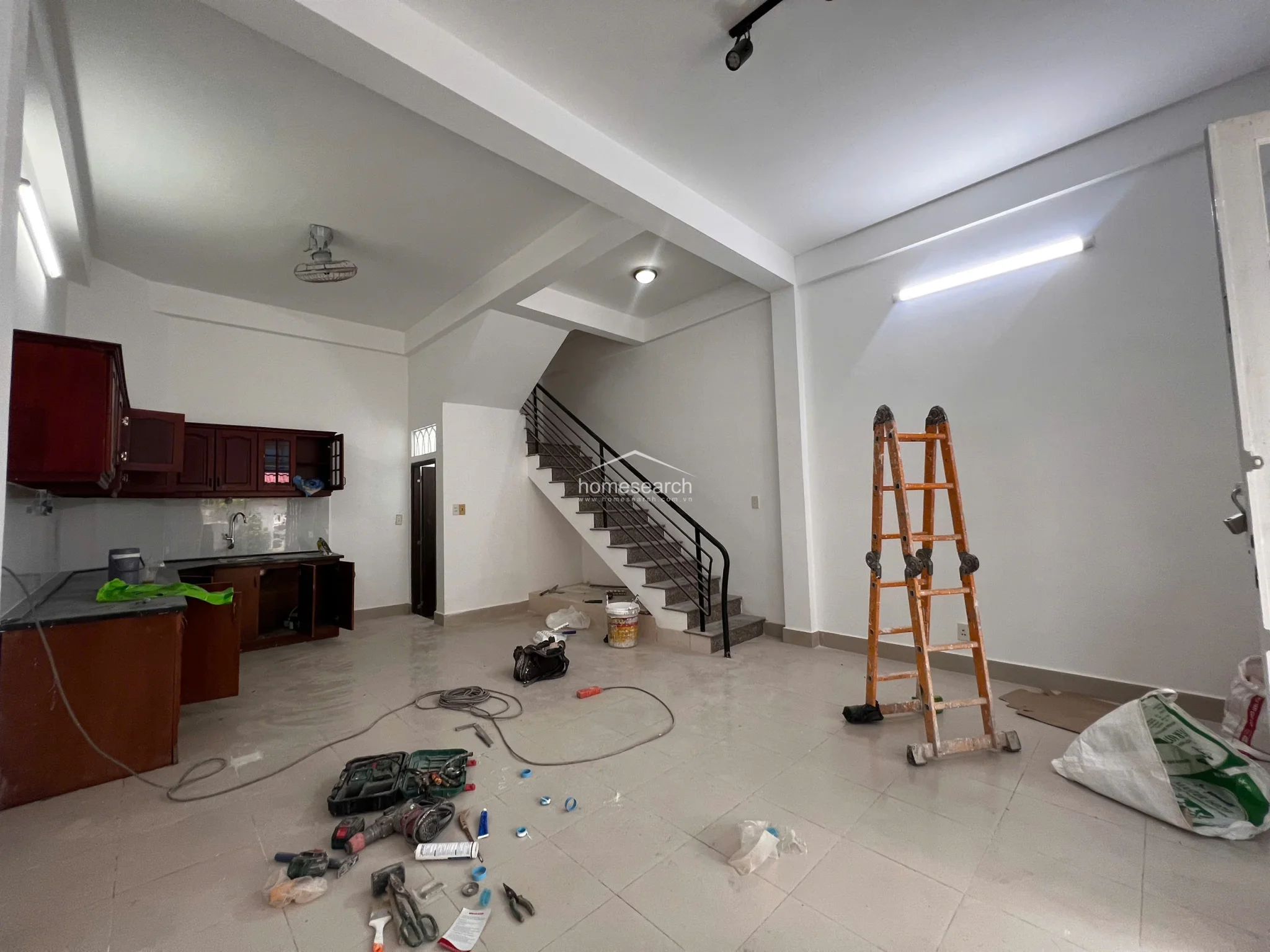 This perspective shows the full depth of the ground floor, including a staircase with a metal railing that leads to the upper levels. The space maintains a consistent aesthetic with light tile floors and white walls, while the presence of construction equipment and a ladder indicates that the property is currently undergoing finishing touches or repairs.