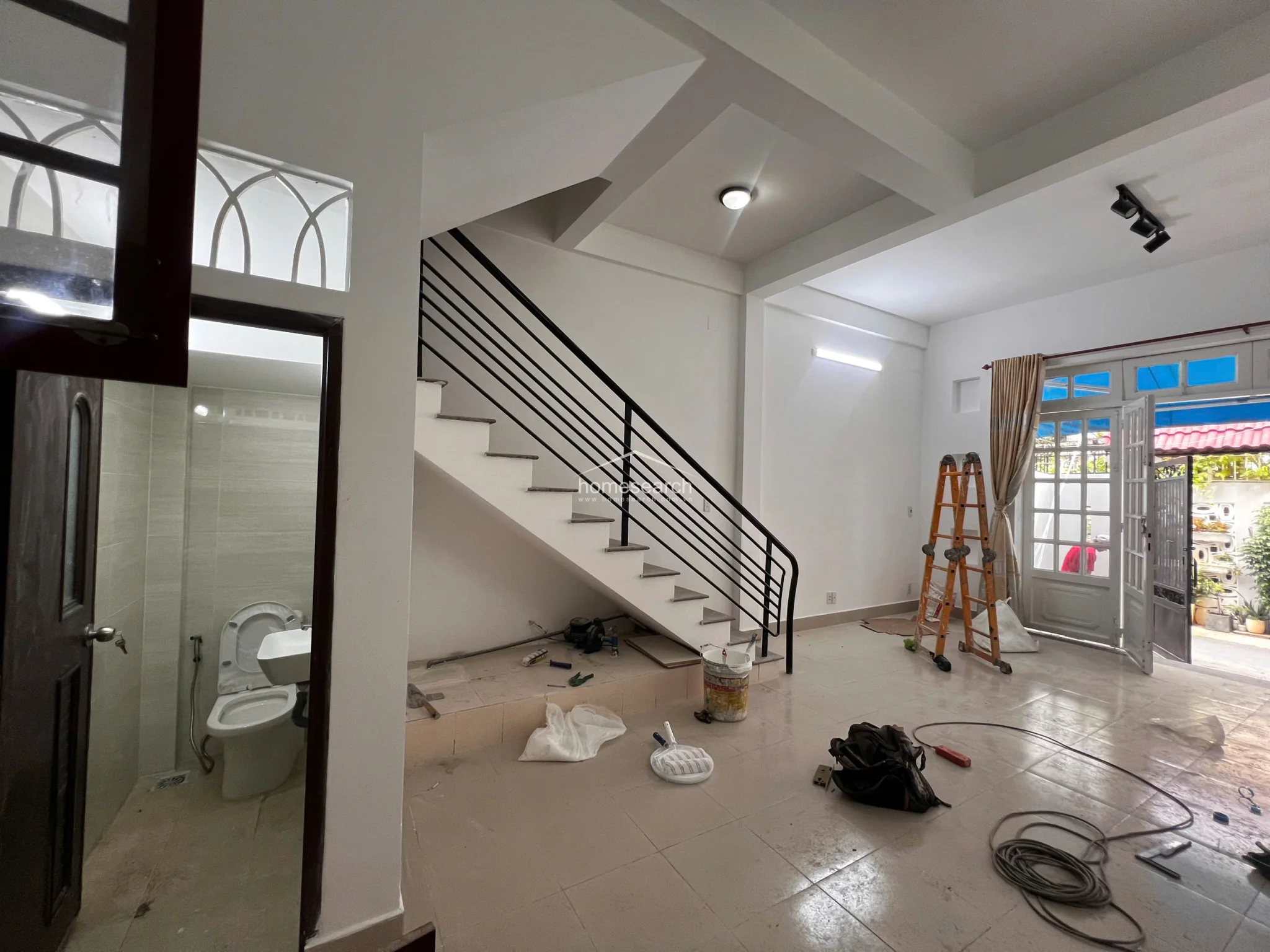 This open-plan living area features light-colored tile flooring and white walls, creating a bright and neutral base currently undergoing minor maintenance. The space includes a staircase with a modern black metal railing and provides direct access to a private outdoor area through a glass-paned door, while a small, functional bathroom is conveniently tucked underneath the stairs.