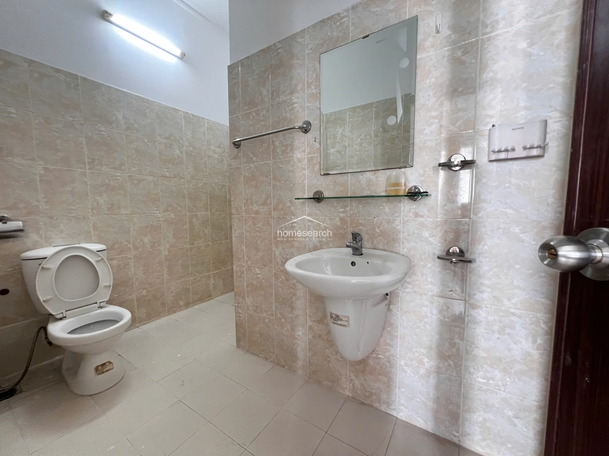 This bathroom features a cohesive design with light, marbled-effect wall and floor tiles that extend throughout the space. It is equipped with a pedestal sink, a wall-mounted mirror with a glass shelf, and a standard toilet, presenting a clean and well-maintained appearance.
