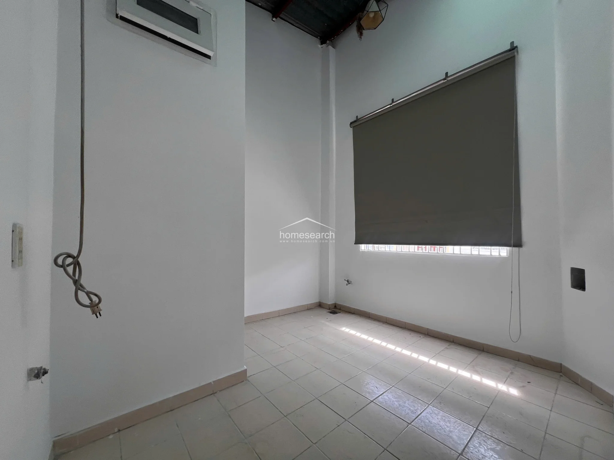 This room features a large window covered by a dark roller blind, providing adjustable natural light and privacy. The space is finished with clean white walls and tile flooring, and includes a ceiling-mounted light fixture and a wall-mounted electrical outlet.