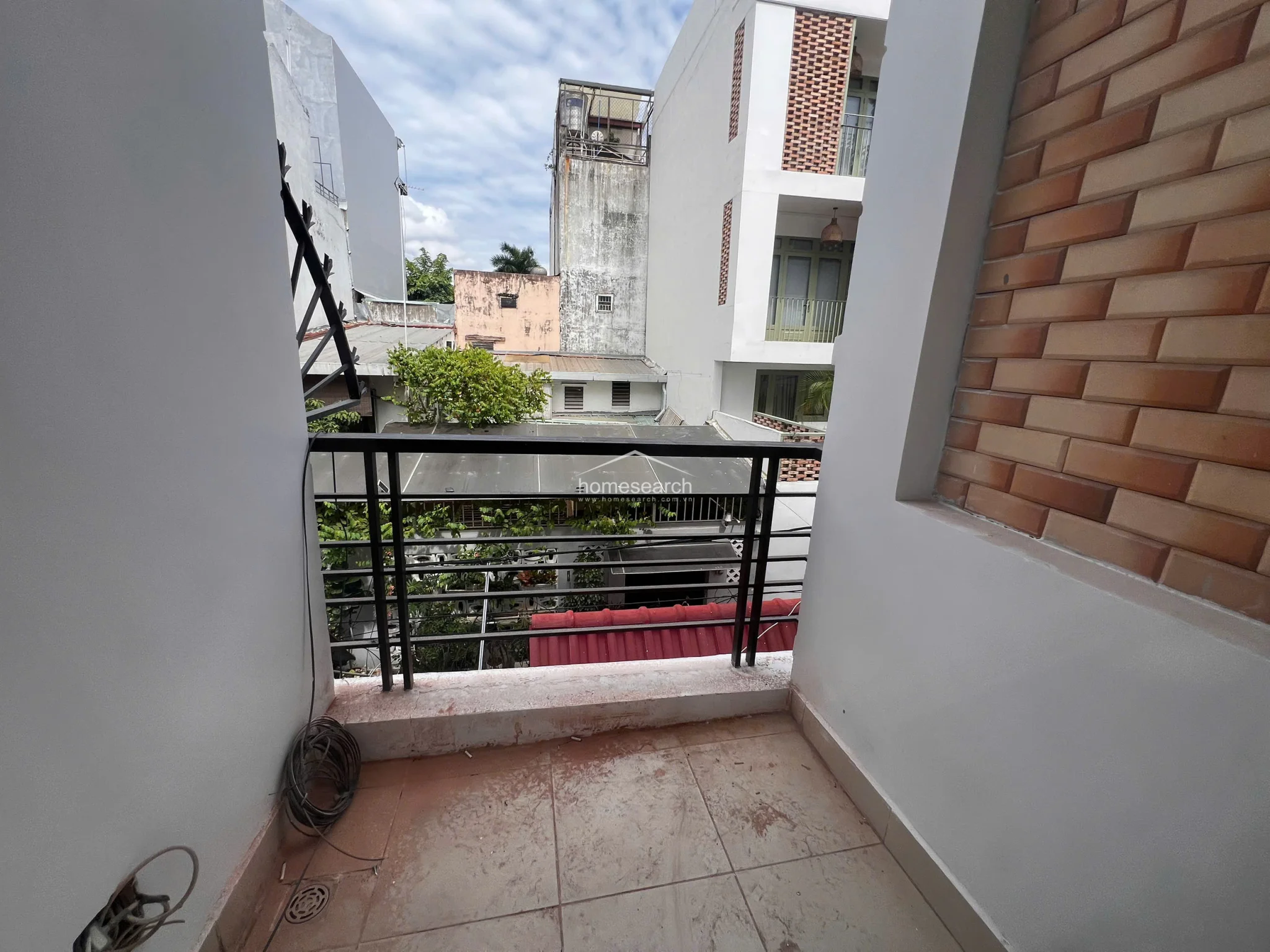 This outdoor balcony features a simple black metal railing and light-colored floor tiles, offering an urban view of neighboring residential buildings. The space is compact and functional, though it shows signs of wear and would benefit from a deep cleaning or minor maintenance.