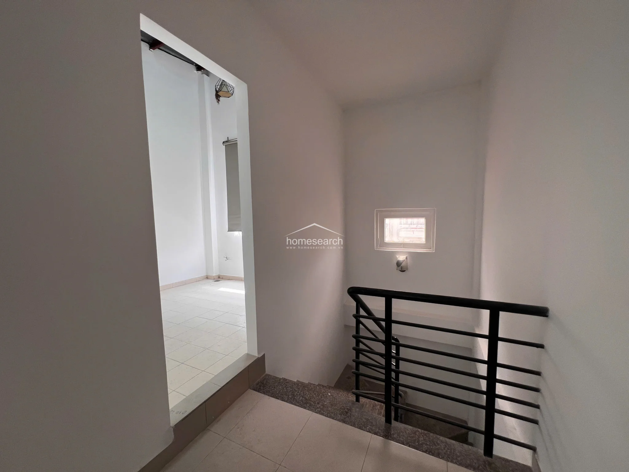 This landing area serves as a central circulation point, featuring neutral tile flooring and a small, high-placed window that introduces natural light into the stairwell. The space connects directly to an adjacent room through an open doorway, maintaining a consistent, minimalist aesthetic with white walls and a simple, functional layout.
