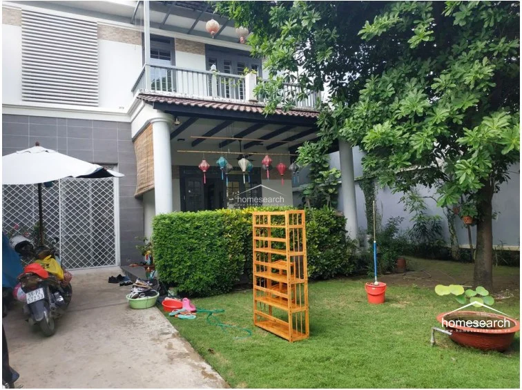 This image shows the exterior of a two-story residence featuring a covered patio, a balcony, and a lush private garden. The architecture includes white columns and a neutral color palette, complemented by decorative hanging lanterns and a well-maintained lawn.
