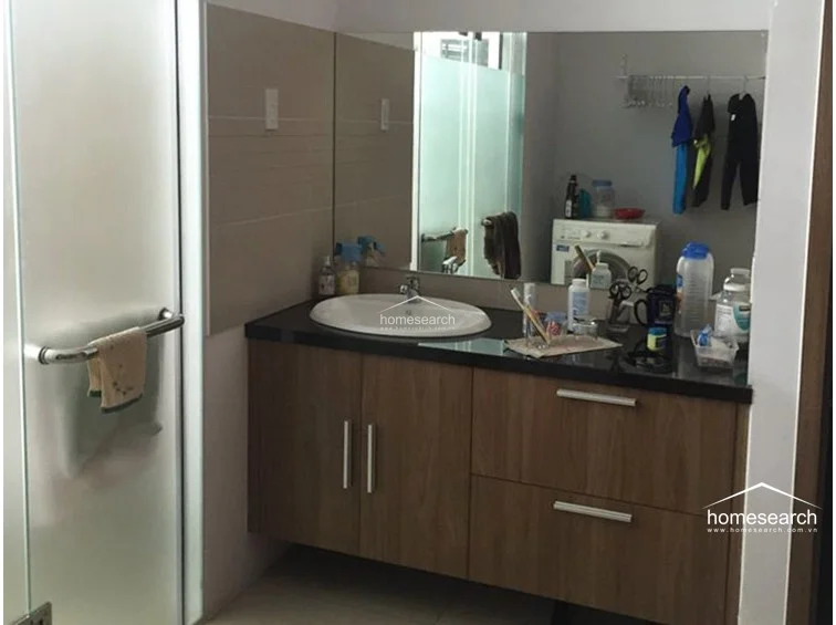 This bathroom features a modern floating vanity with a dark stone countertop and wood-grain cabinetry, providing a clean and contemporary look. The space includes a glass-enclosed shower area and a large mirror, though the room is currently cluttered with personal items and a washing machine, suggesting a multi-functional utility space.