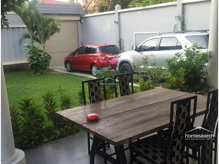 This image focuses on the covered patio area, which serves as an ideal spot for outdoor dining with a large wooden table and patterned chairs. The space overlooks the driveway and garden, creating a seamless flow between the home's interior and the parking area.