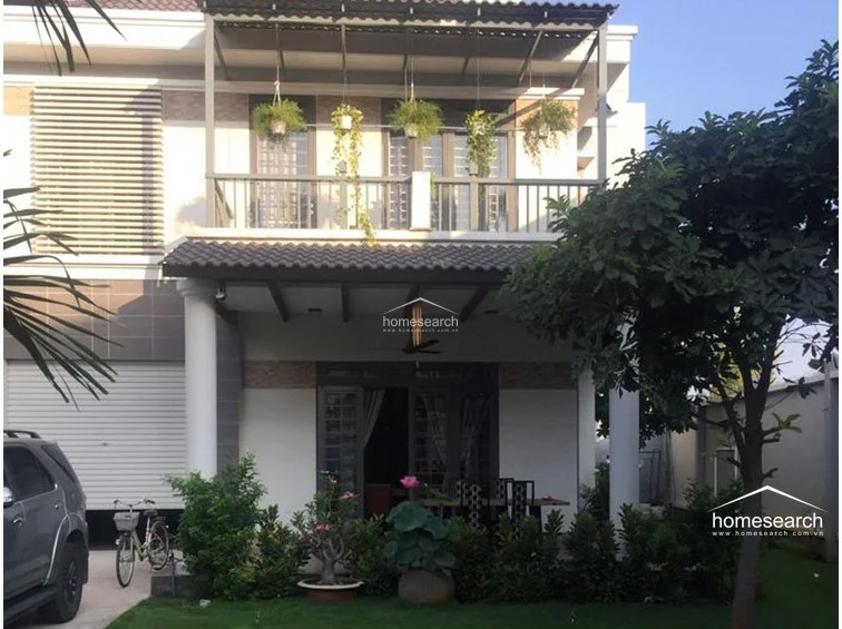 The exterior of the property showcases a contemporary two-story design with a spacious covered ground-floor patio and a balcony above, both adorned with hanging plants. The architecture includes clean lines, white pillars, and a tiled roof, complemented by a well-maintained lawn and mature trees in the front garden.