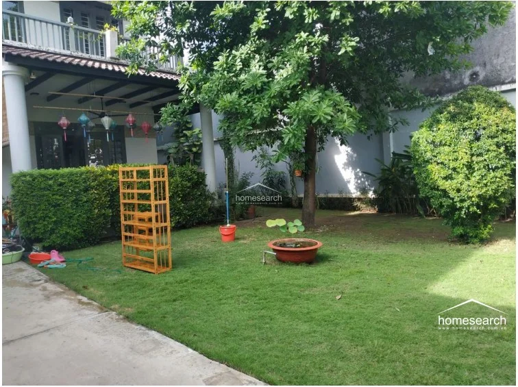 This private garden area offers a tranquil retreat with a lush green lawn, mature trees, and neatly trimmed hedges providing natural privacy. The space is functional and well-maintained, featuring a decorative water feature and a portable wooden shelving unit, creating a peaceful outdoor environment.