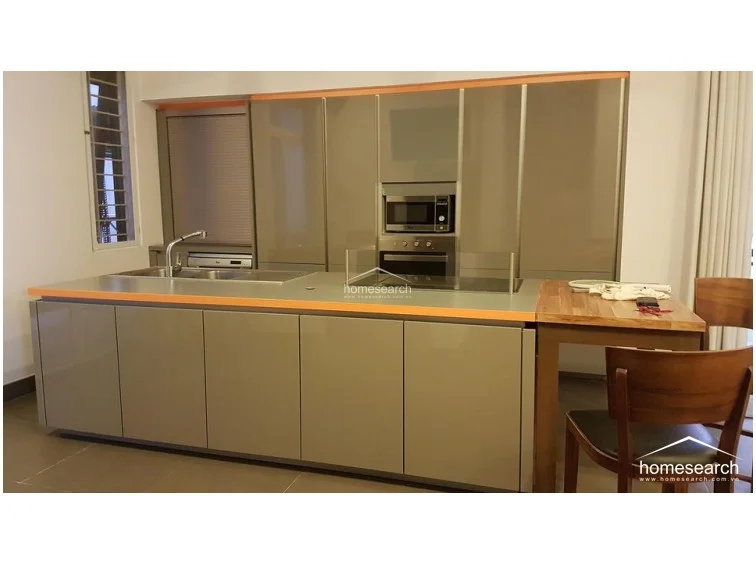 The kitchen features a sleek, high-end design with glossy grey cabinetry, a built-in oven and microwave, and a large central island with an integrated sink and cooktop. An attached wooden breakfast bar extends the workspace, while the overall layout emphasizes clean, modern lines and efficient storage solutions.