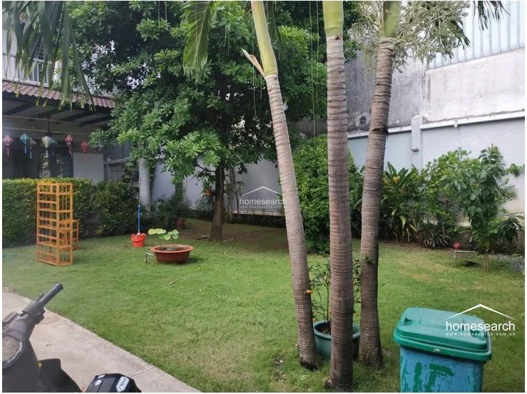 This exterior view showcases a private garden area with mature palm trees and lush landscaping, offering a tranquil outdoor retreat. The property features a covered patio with decorative lanterns and a balcony above, indicating a well-designed architectural layout that blends indoor and outdoor living.