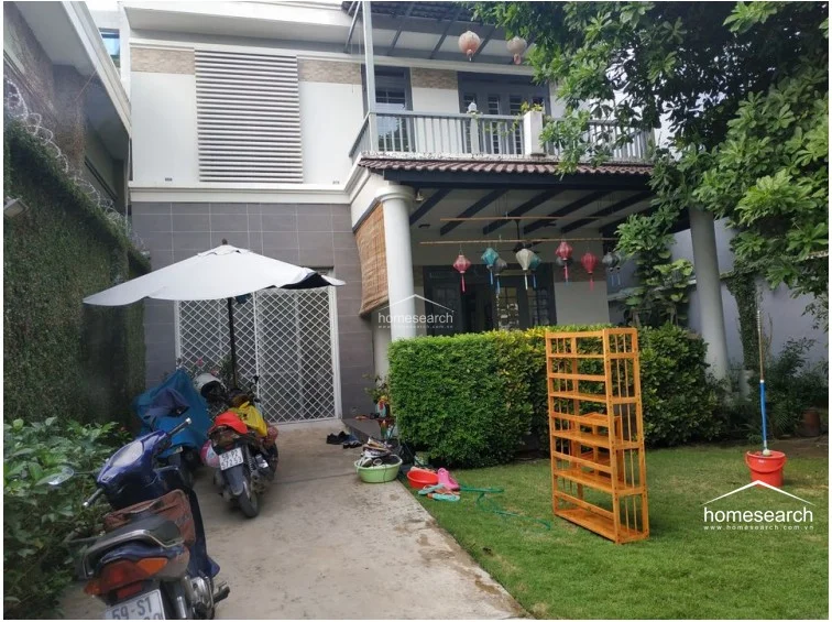 This perspective of the home's exterior emphasizes the connection between the paved driveway and the green garden space, framed by mature trees. The architectural design includes a distinct balcony with a metal railing and a covered ground-level patio, suggesting a comfortable and practical flow for family living.
