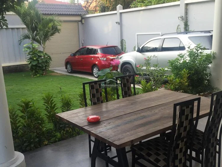 This outdoor dining area is situated on a covered patio, featuring a rustic wooden table and patterned chairs that overlook the front yard. The space offers a clear view of the driveway and garden, providing a functional and relaxed setting for outdoor entertaining.