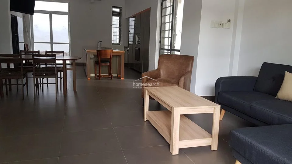 This open-concept living and dining area features a modern, minimalist aesthetic with large-format grey floor tiles and crisp white walls. The space flows seamlessly into a contemporary kitchen, with ample natural light provided by large glass doors and windows that offer a view of the outdoor greenery.