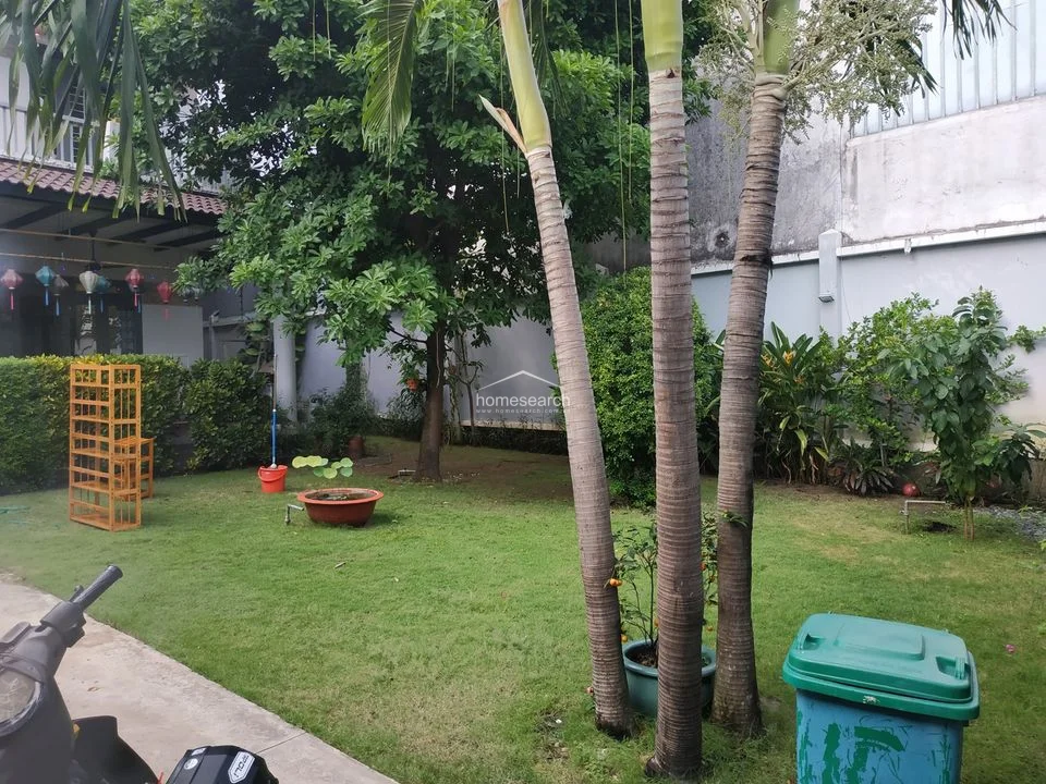This shot captures the expansive private garden, which features mature trees, palm plants, and a manicured lawn. The space is enclosed by a high perimeter wall, ensuring privacy and a tranquil atmosphere for outdoor relaxation.