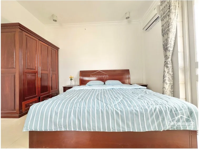 The bedroom is anchored by a large, traditional wooden wardrobe and a matching bed frame, creating a warm and classic aesthetic. A wall-mounted AC unit and a window with patterned curtains ensure comfort and natural light, while the neutral tile flooring keeps the room feeling airy and spacious.