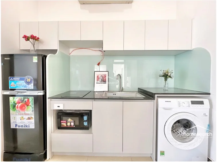 This compact kitchenette is efficiently laid out with white cabinetry, a light-green glass backsplash, and a dark stone countertop. It is equipped with a Funiki refrigerator, a built-in microwave, and a Samsung washing machine, making it a highly functional space for daily needs. The area is well-lit and maintained, with clean surfaces and modern fixtures.