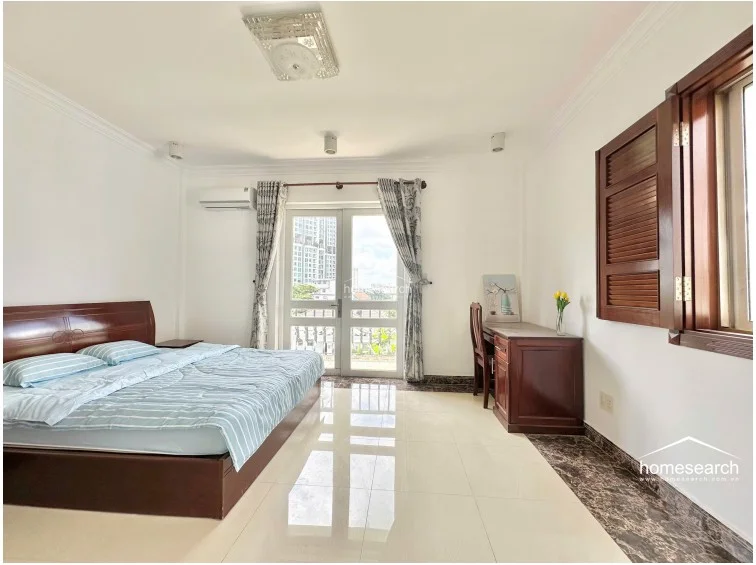 This spacious bedroom features light-colored tile flooring with a decorative dark border and is illuminated by natural light from a glass-paned door that opens onto a balcony. The room is furnished with a dark wood bed frame and a matching desk, complemented by a wall-mounted air conditioning unit and a ceiling-mounted light fixture. The walls are finished in a clean white, creating a bright and well-maintained atmosphere.