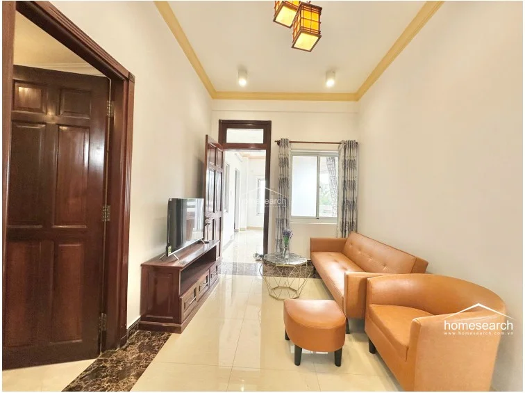 The living room layout flows directly into the adjacent kitchen and provides a clear view into the bedroom area through an open doorway. It is furnished with a vibrant orange sofa and matching ottoman, a geometric marble-top coffee table, and a wooden media console, all set against a clean, minimalist backdrop.