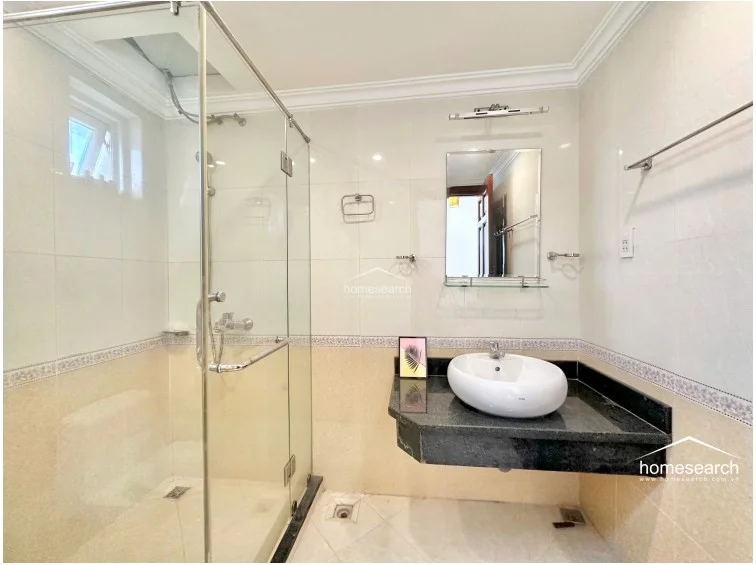 This bathroom features a walk-in glass shower enclosure, a white vessel sink set on a dark granite floating vanity, and a modern toilet. The walls are tiled in a neutral two-tone scheme with a decorative border, and a small window provides natural ventilation and light. The space appears to be in good condition with clean, functional fixtures.