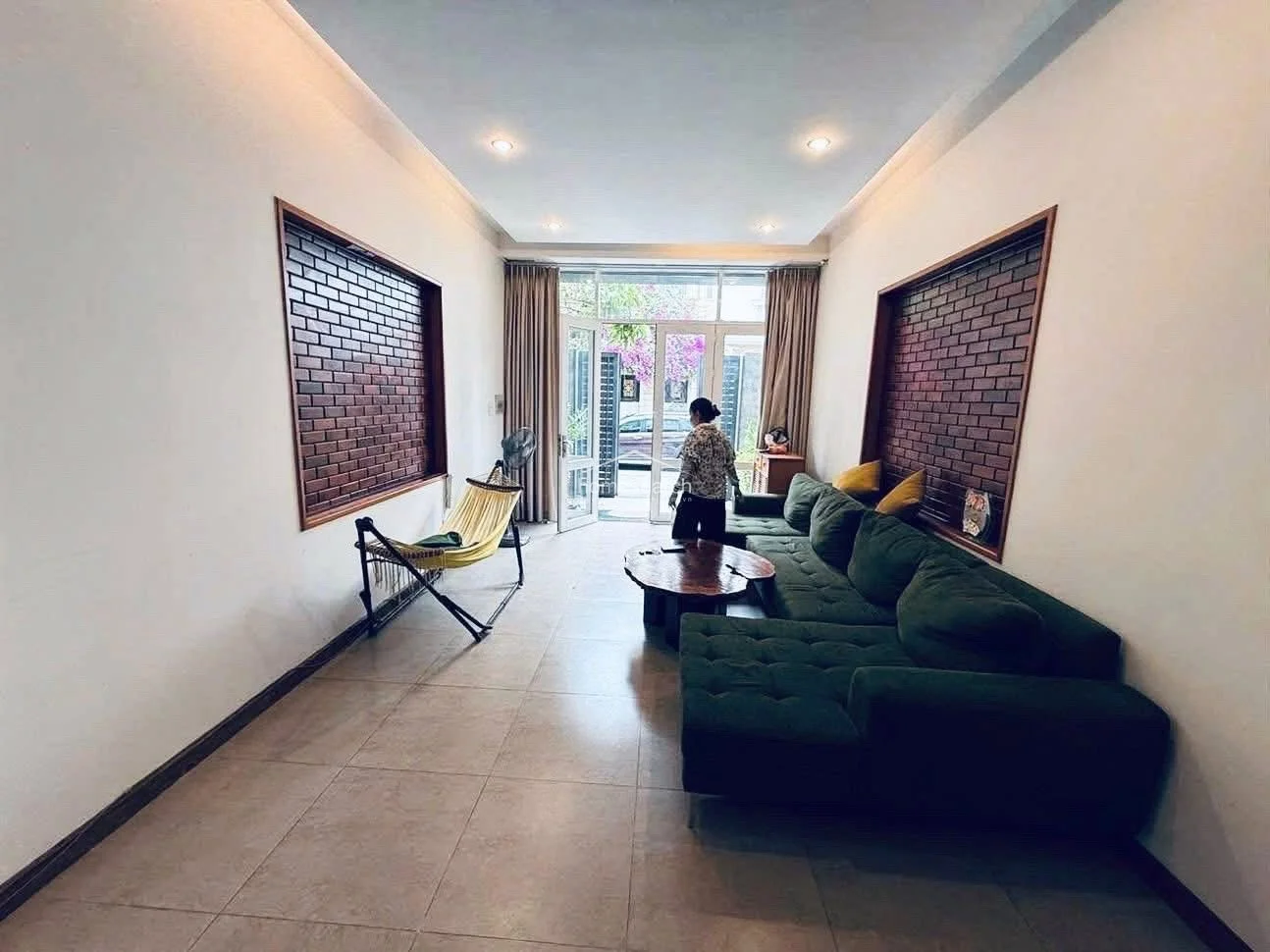 This living room features a modern, open-plan layout with large glass doors that provide excellent natural light and a view of the exterior. The space is anchored by a large, dark green L-shaped sofa and a unique natural-edge wooden coffee table, complemented by decorative brick-accented walls and neutral tile flooring.