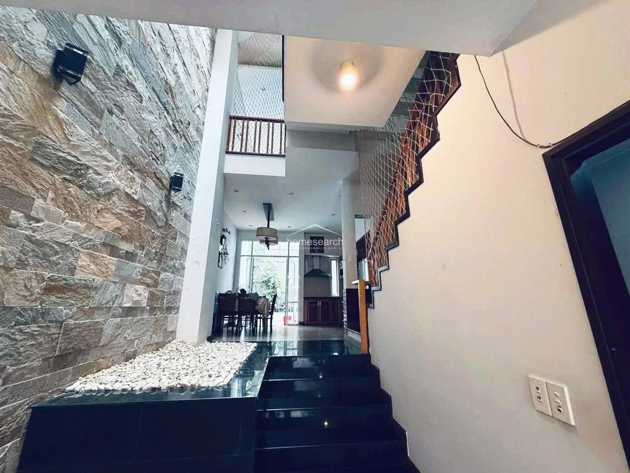 This transitional area showcases a striking architectural feature with a tall, textured stone wall that adds depth and character to the home. The space includes a small indoor garden bed with white stones and a staircase leading to the upper levels, which is secured by a safety net for an open, airy feel.