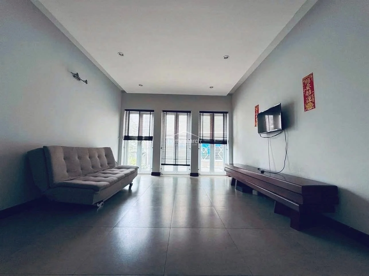This secondary living or family room is minimalist in design, featuring a neutral grey sofa bed and a long, low-profile wooden media console. It benefits from natural light streaming through three vertical windows fitted with dark blinds, set against clean, light-colored walls and matching tile floors.