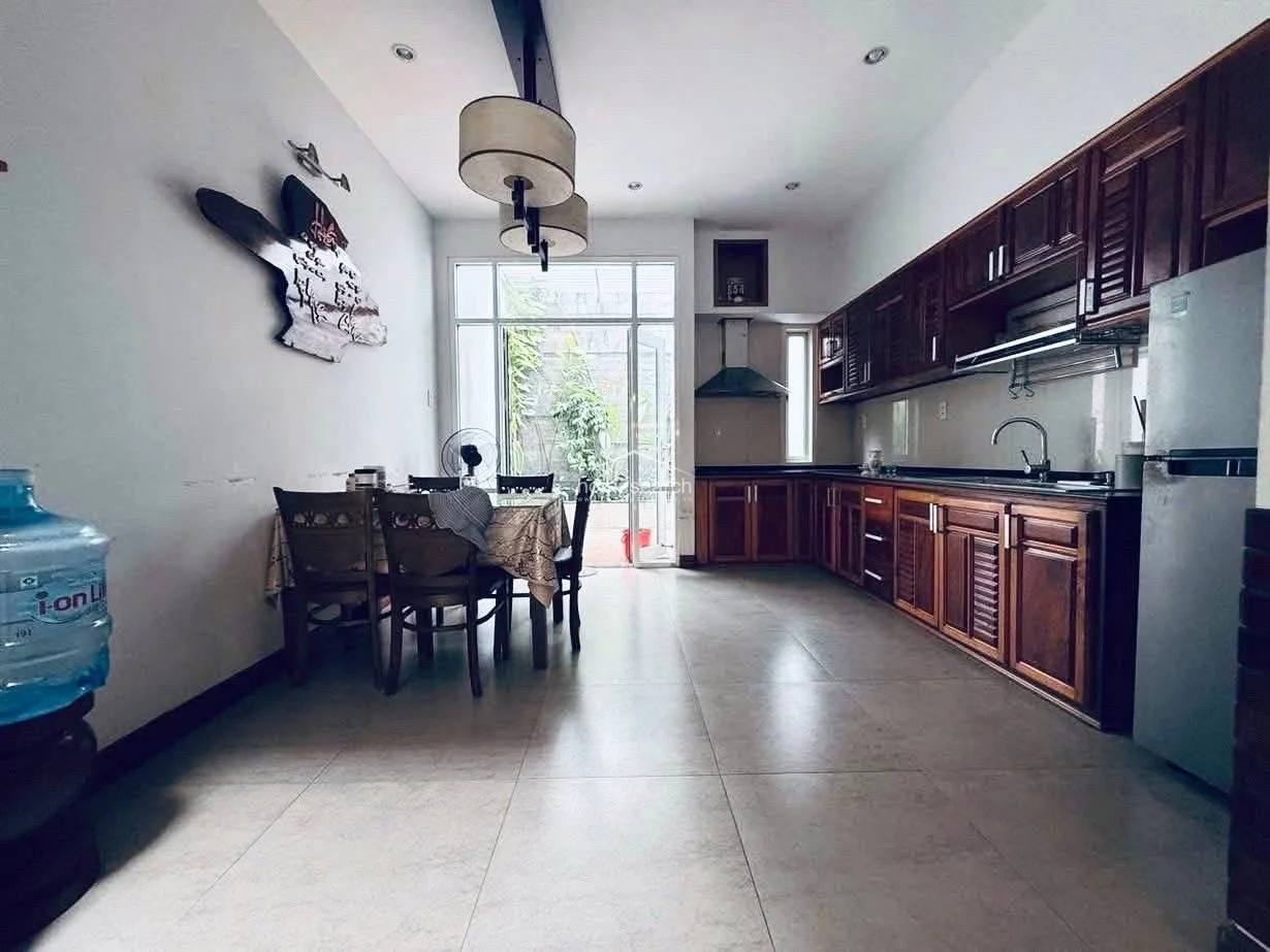 The kitchen and dining area is designed for functionality, featuring a large wooden dining set and an extensive L-shaped kitchen with rich, dark wood cabinetry. The space is well-equipped with a stainless steel range hood, a full-sized refrigerator, and large glass doors that open to an outdoor area, ensuring a bright and ventilated environment.