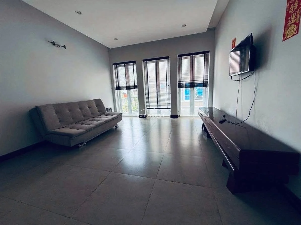 This minimalist living area features large windows with dark blinds that allow for ample natural light and a view of the exterior. The space is finished with neutral grey walls, large-format floor tiles, and includes a modern grey sofa and a long, dark wood media console.