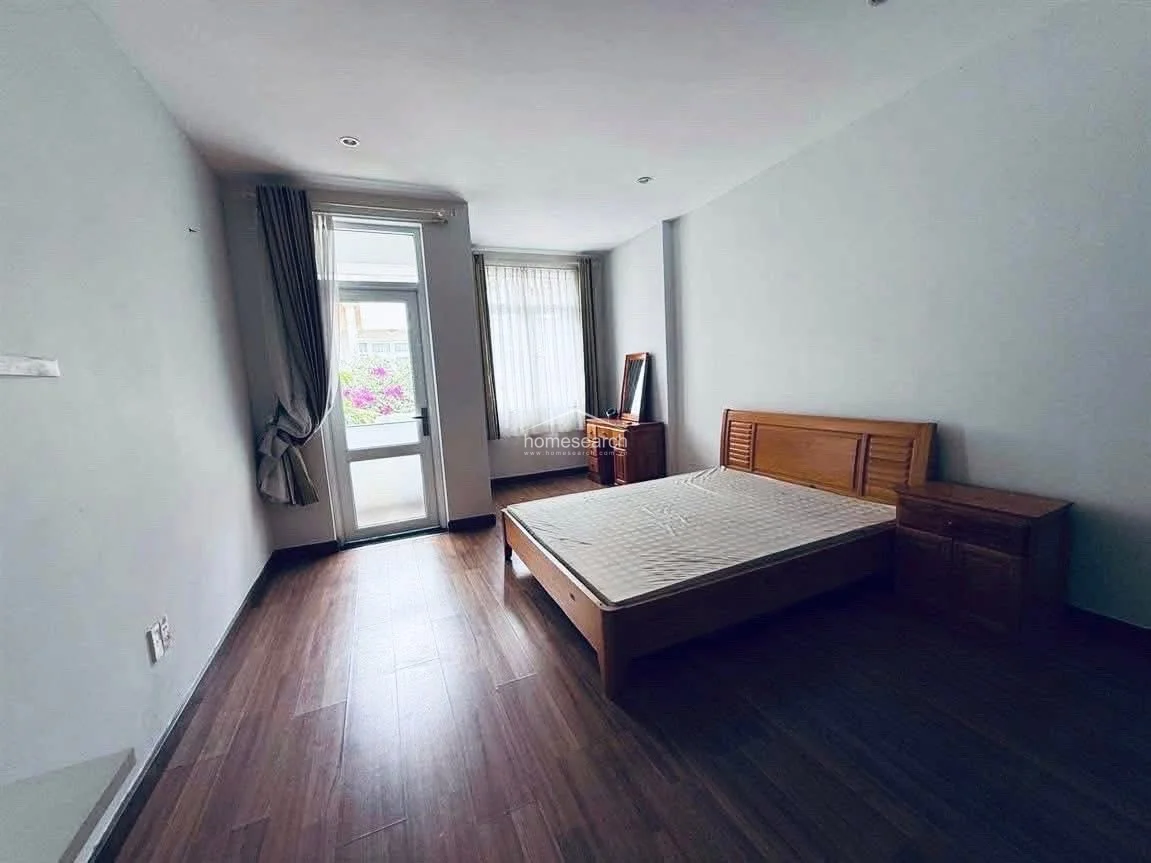 This bedroom features warm wood-look flooring and a matching wooden bed frame and vanity, creating a cohesive and inviting aesthetic. A glass door provides access to an outdoor balcony area, while sheer curtains allow for soft, natural light to filter into the room.