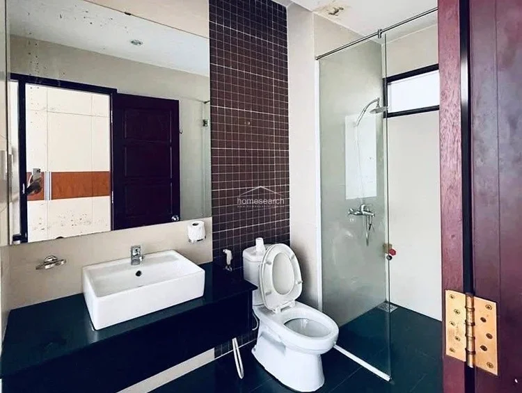 The bathroom is designed with a modern aesthetic, featuring a dark mosaic tile accent wall and a sleek black vanity countertop. It includes a glass-enclosed walk-in shower, a contemporary white vessel sink, and a large wall-mounted mirror that enhances the sense of space.