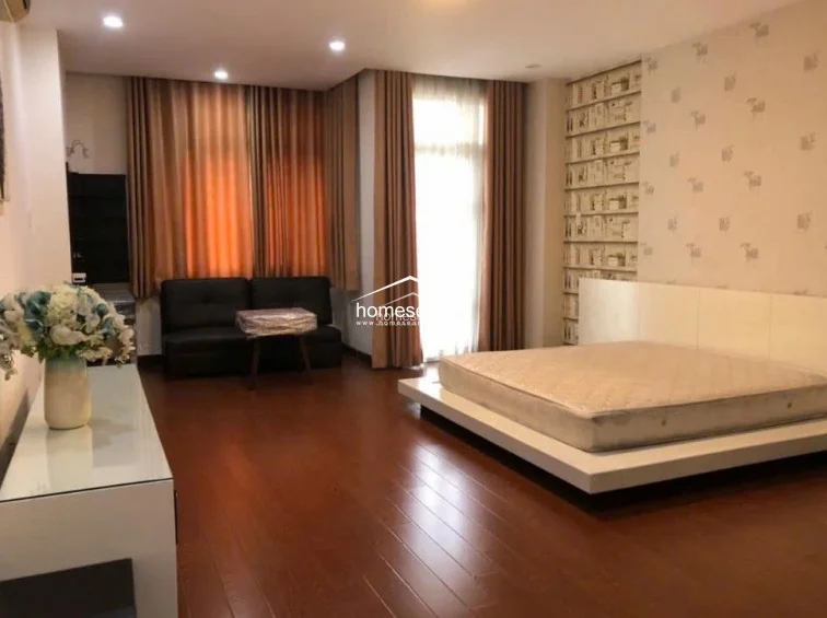 This spacious bedroom features rich wooden flooring and a platform bed, creating a contemporary and serene sleeping environment. A large window draped with floor-to-ceiling curtains allows for significant natural light, while the wall behind the bed is accented with patterned wallpaper. The room is well-maintained, offering a clean, uncluttered layout with a small seating area and a minimalist white console table.