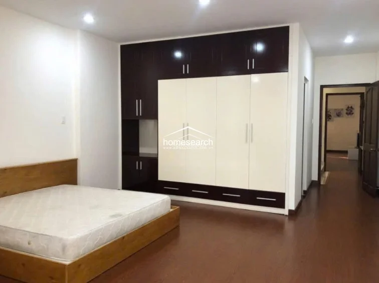 This spacious bedroom features warm wood-tone flooring and crisp white walls, creating a clean and minimalist aesthetic. It includes a large, floor-to-ceiling built-in wardrobe with a modern dark wood and cream-colored finish, providing extensive storage, and connects directly to a hallway leading to other areas of the home.