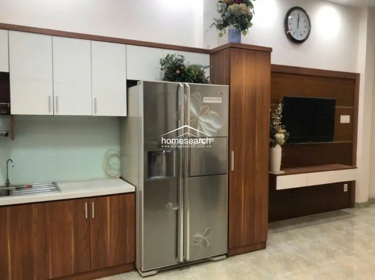 The kitchen is well-equipped with a large stainless steel double-door refrigerator, sleek white upper cabinets, and warm wood-toned lower cabinetry. A built-in media console on the adjacent wall continues the wood theme, creating a cohesive design language that integrates the kitchen seamlessly into the living environment.