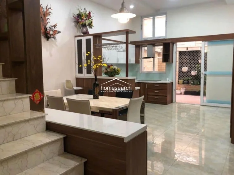 This open-concept kitchen and dining area features a modern, warm aesthetic with wood-toned cabinetry and a light-colored dining set. The space flows seamlessly into a rear patio through a large sliding glass door, which provides ample natural light and a view of the outdoor greenery. The flooring consists of large, polished light-colored tiles, and the area is well-maintained with integrated kitchen appliances and a stylish pendant light above the dining table.
