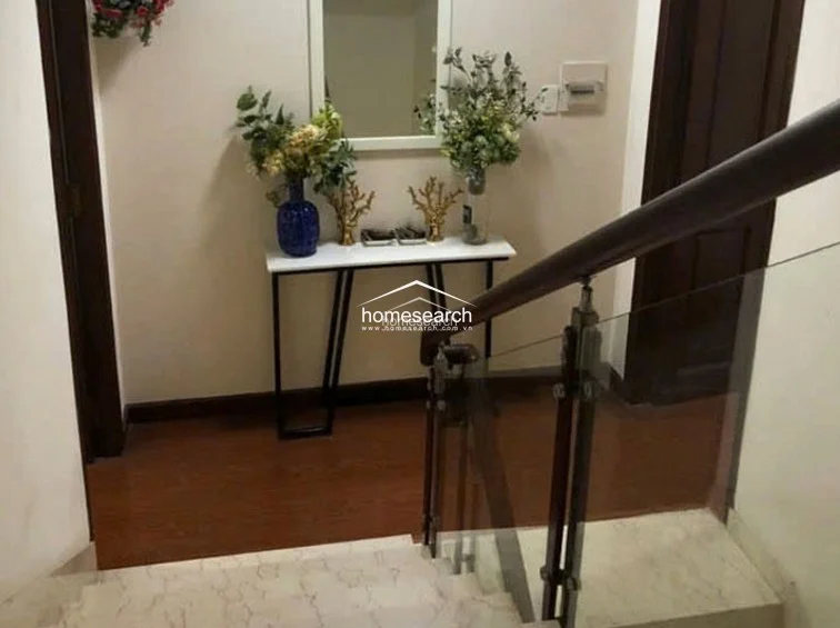 This landing area serves as a stylish transition space, featuring a modern console table topped with decorative floral arrangements and a wall-mounted mirror. The flooring transitions from polished marble on the stairs to warm-toned wood, complemented by a sleek glass and wood staircase railing. The area is well-lit and thoughtfully decorated, creating a sophisticated and welcoming focal point within the home.